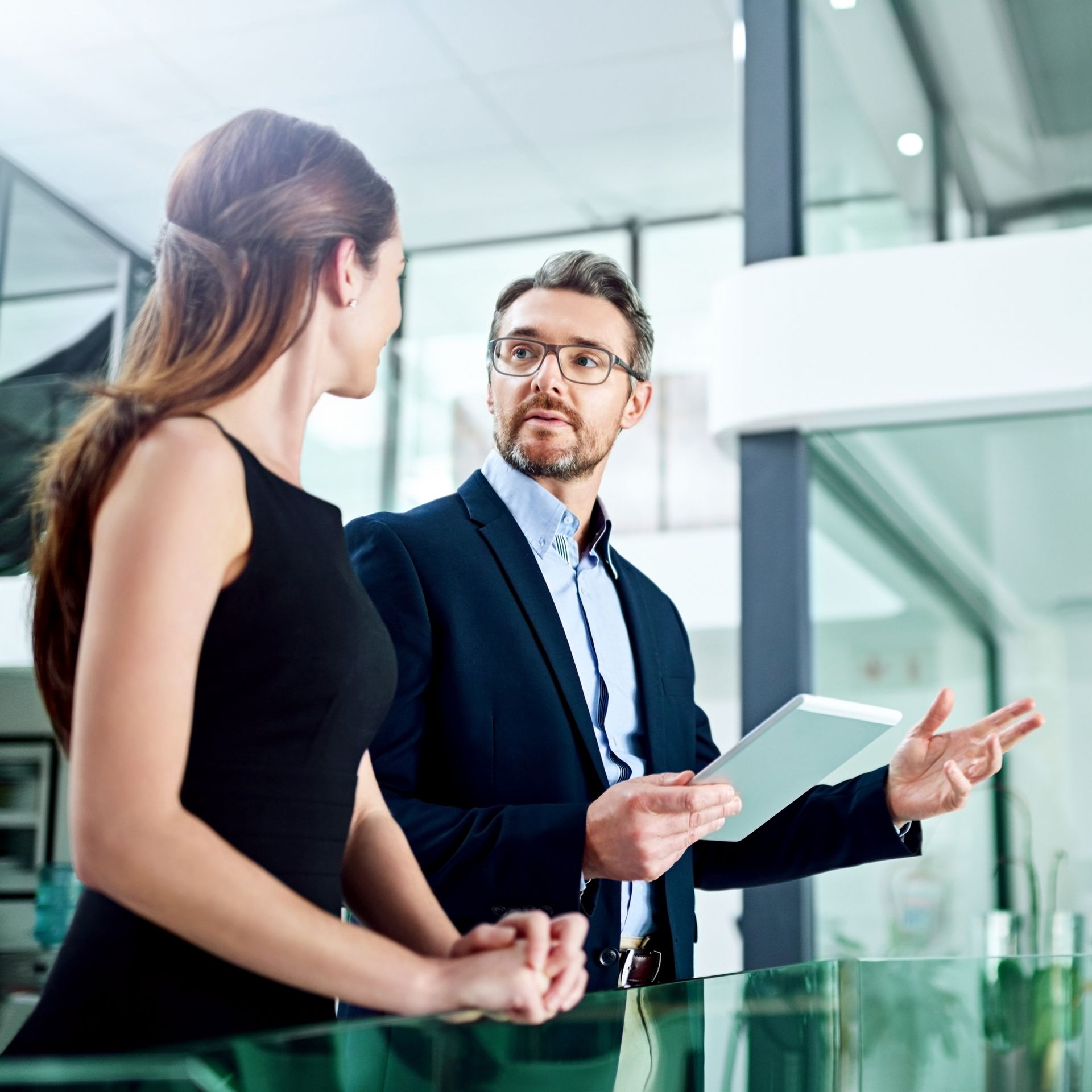 Man in suit gestures, holding tablet, talking to woman in black dress in modern office.