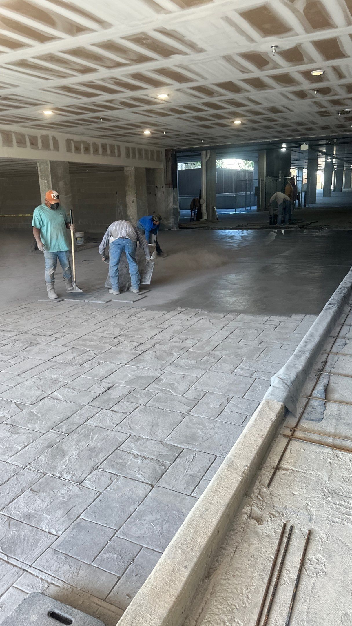 Construction workers pouring concrete on a patterned surface, under a partially finished ceiling, inside a building.