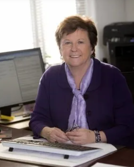 Woman in purple sweater, seated at a desk, smiling, holding glasses, with a computer and printer in the background.
