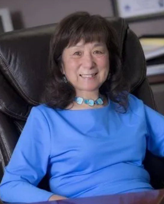 Woman seated in a leather office chair, wearing a blue shirt and turquoise necklace, smiling.