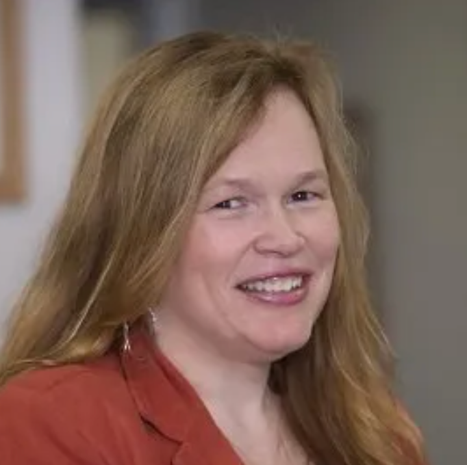 Woman with long blonde hair, wearing a reddish blazer, smiles.