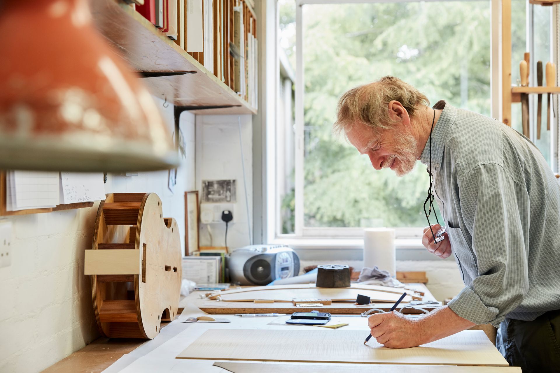 Man drawing at a desk in a workshop, looking at his work. Window and shelves in the background.