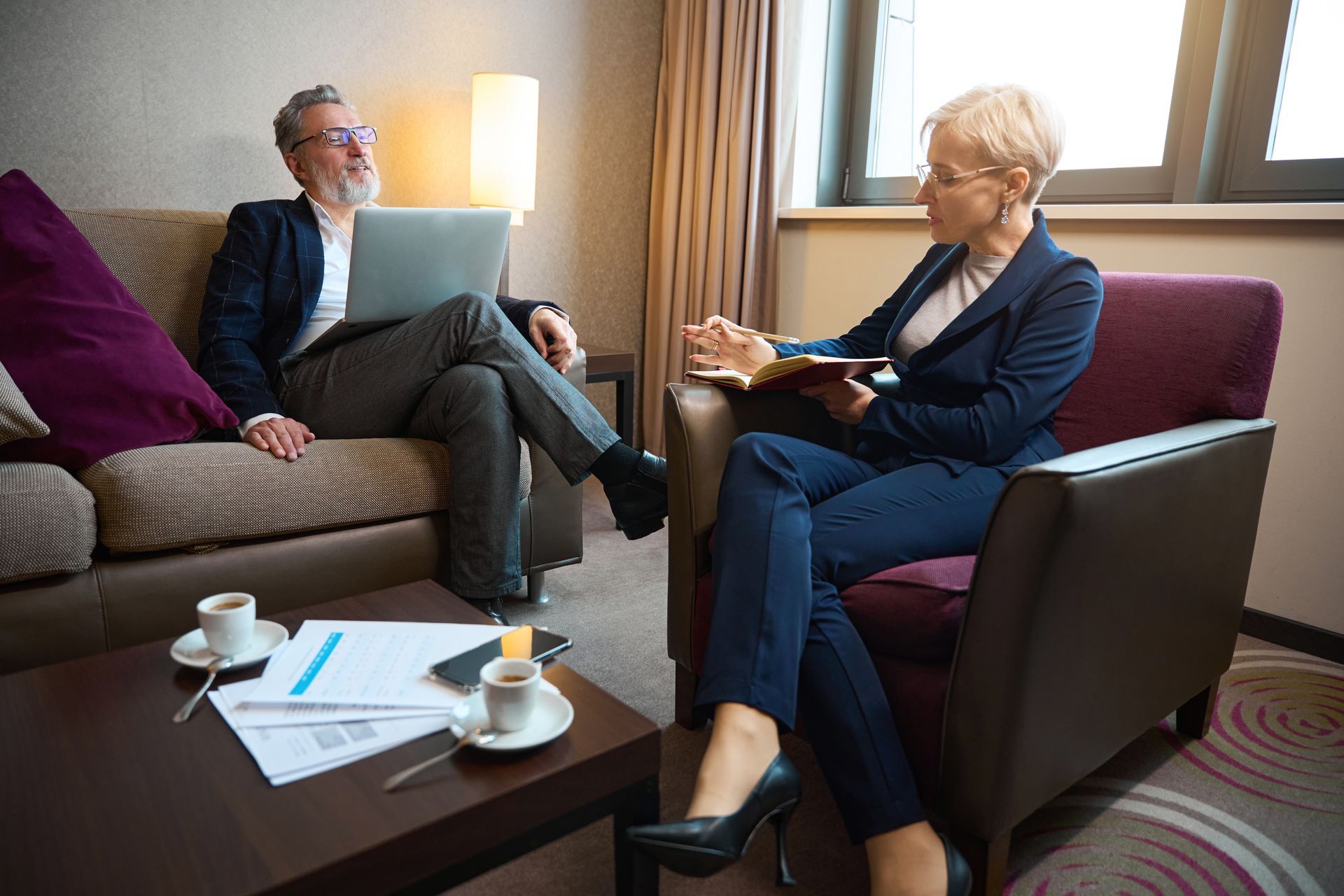 Two people in business attire in a hotel room, one with laptop on sofa, other in armchair with notebook, coffee on table.