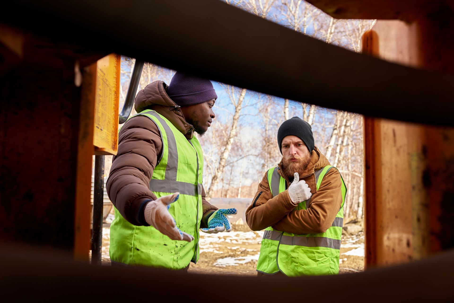 Two workers in safety vests are talking outdoors, one gesturing, the other listening.