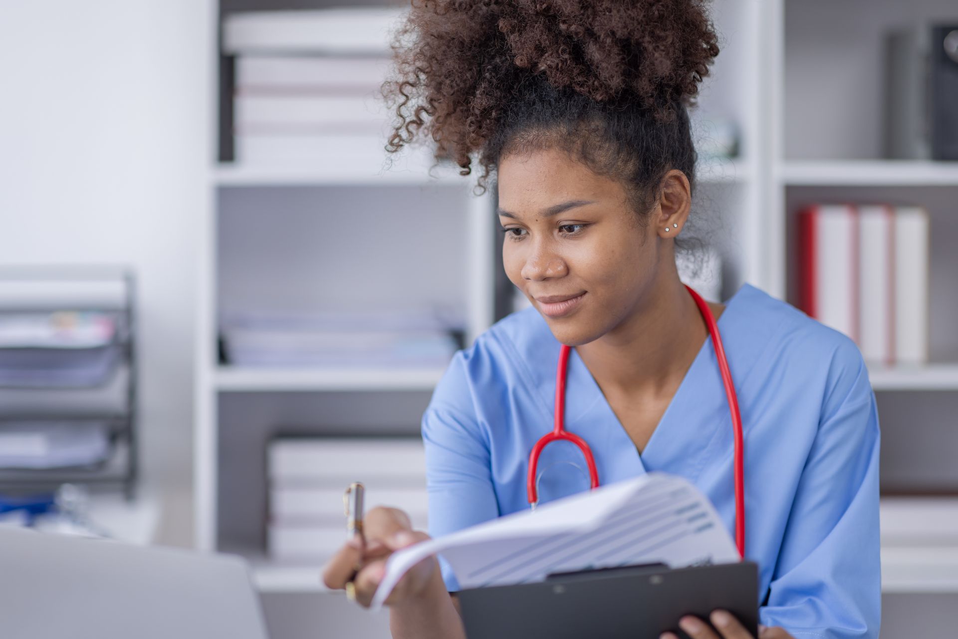 Medical professional reviewing documents, holding a pen, and looking at a laptop with a bookshelf background.