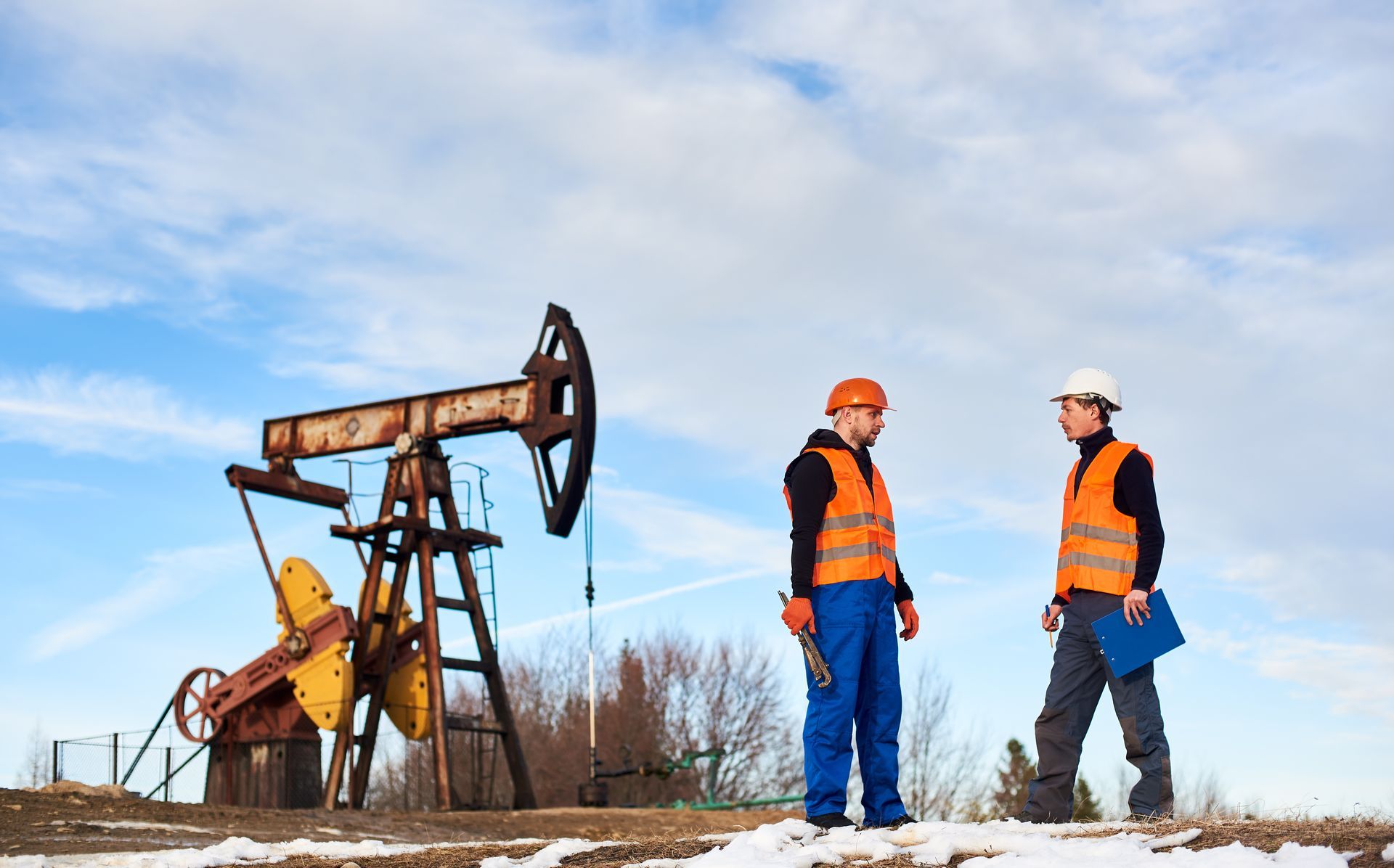 Two workers in safety vests stand near an oil pump in a snowy field under a blue sky.