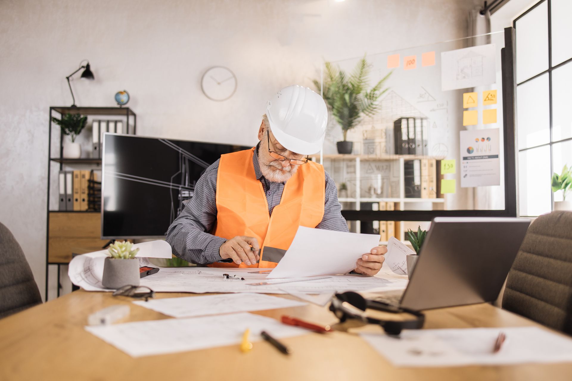 Engineer in safety vest and hard hat reviewing blueprints at a desk with a laptop and office supplies.