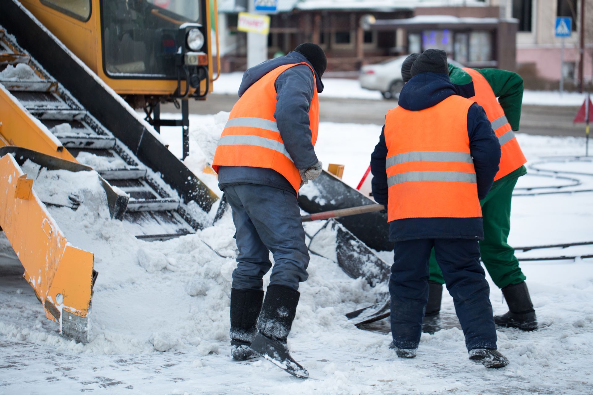 Workers in orange vests shovel snow next to yellow snow removal equipment on a snowy street.