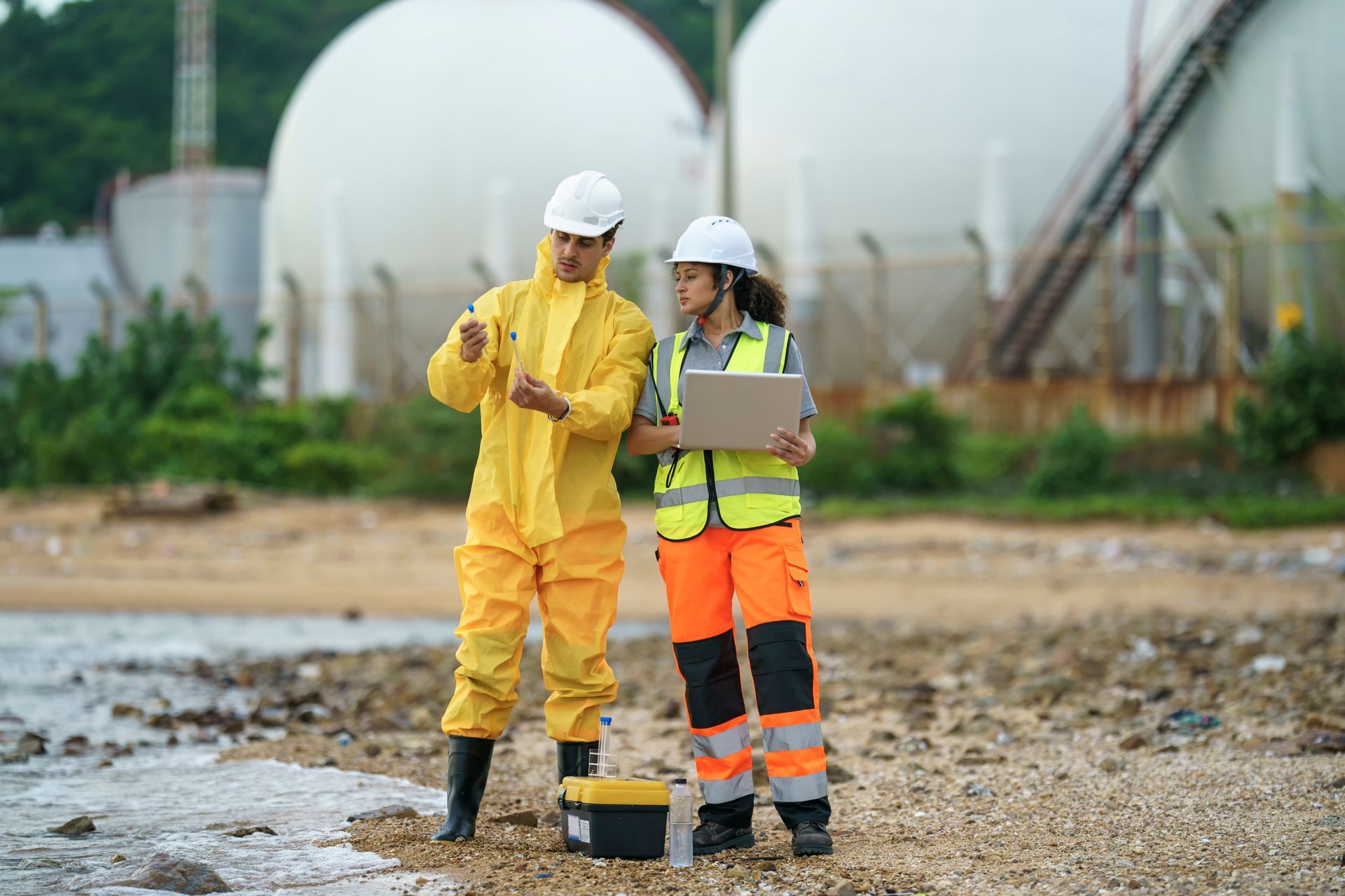 Two people in protective gear examine water near storage tanks. One points while the other holds a laptop.