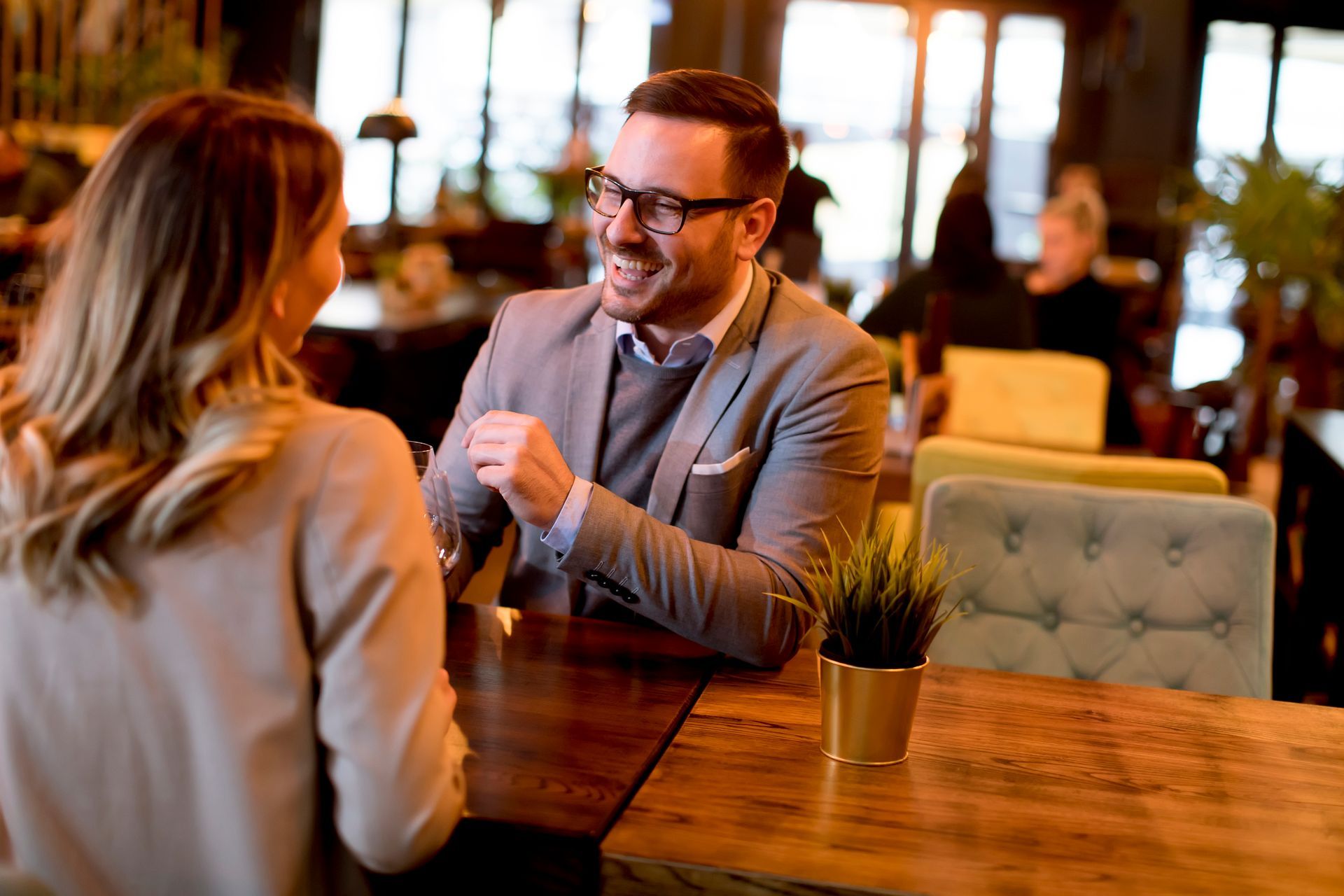 Man and woman smiling and talking at a restaurant table. Man in gray suit and glasses, potted plant.