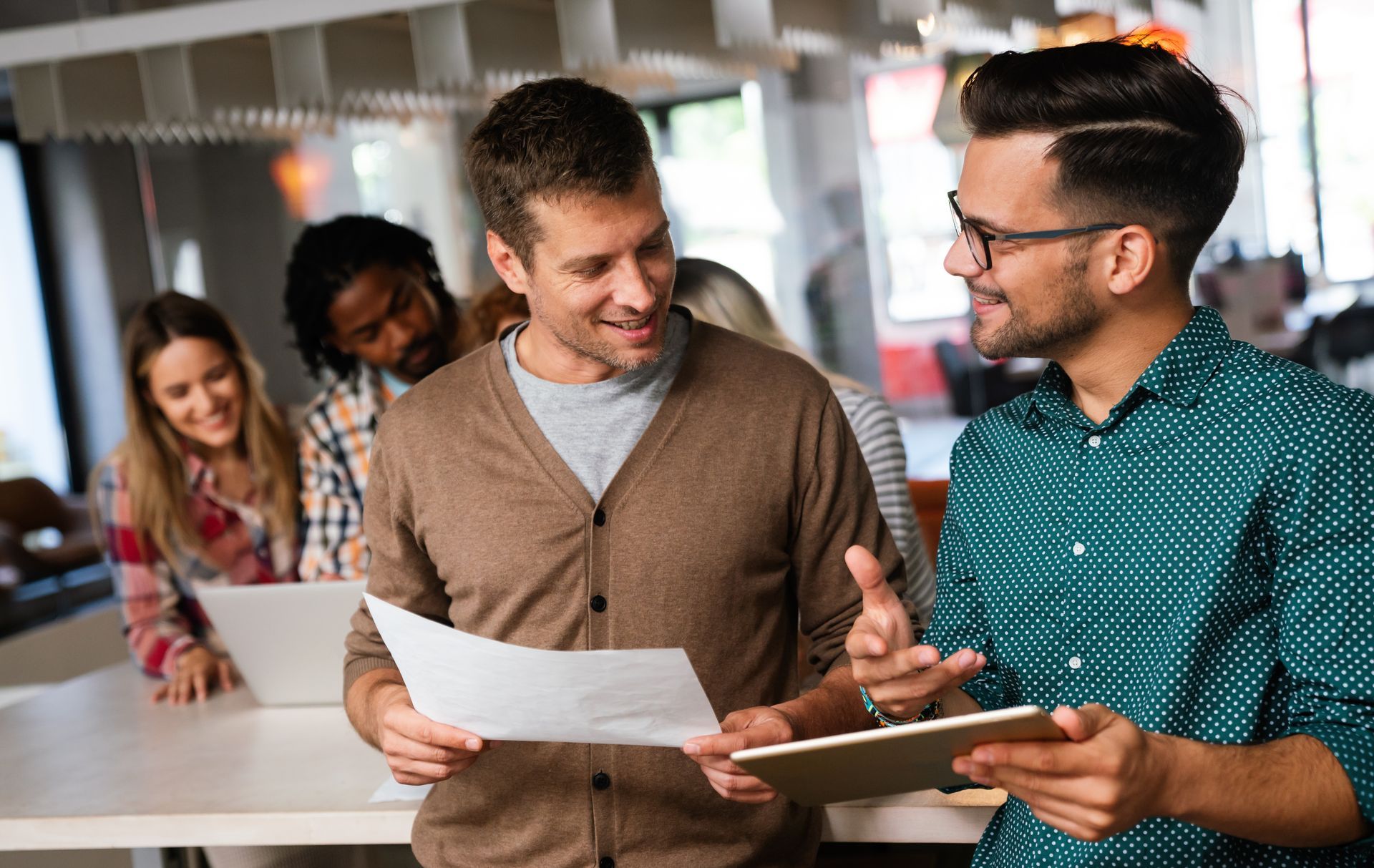 Two men discussing documents in an office, other people in the background working.