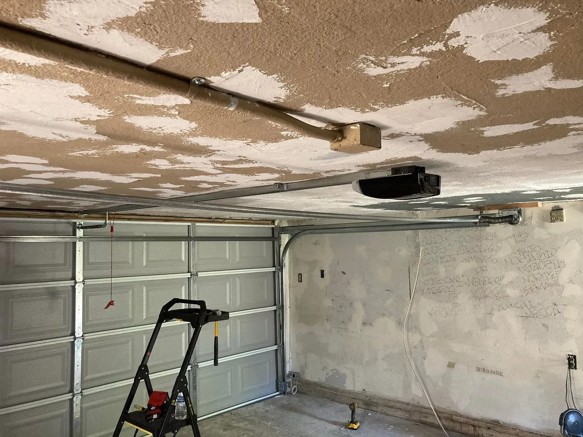 Partially finished garage interior with open door, ladder, and ceiling fixtures on a patchy ceiling