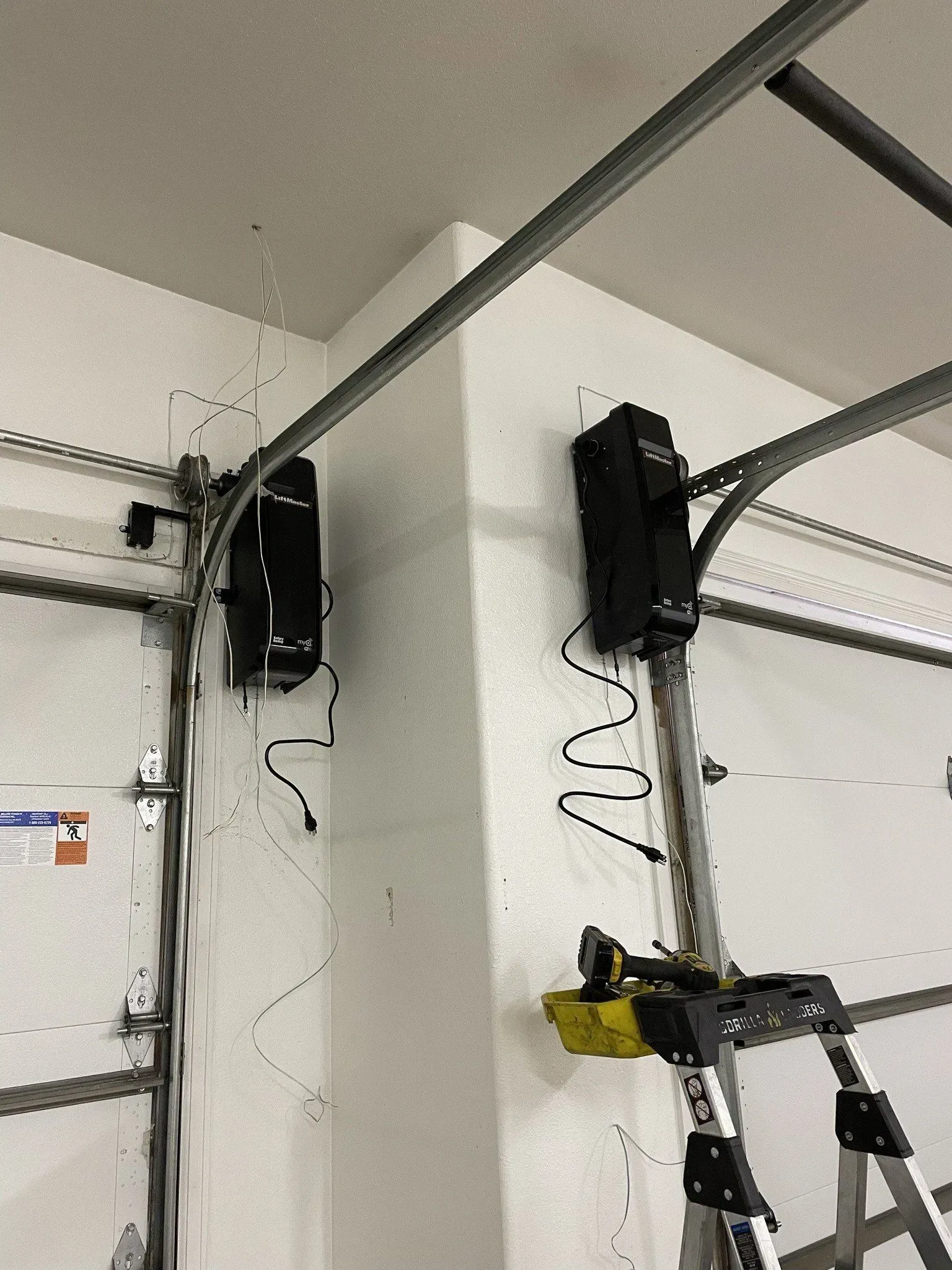 Garage ceiling with two black garage door openers and metal tracks; a ladder leans against one opener.