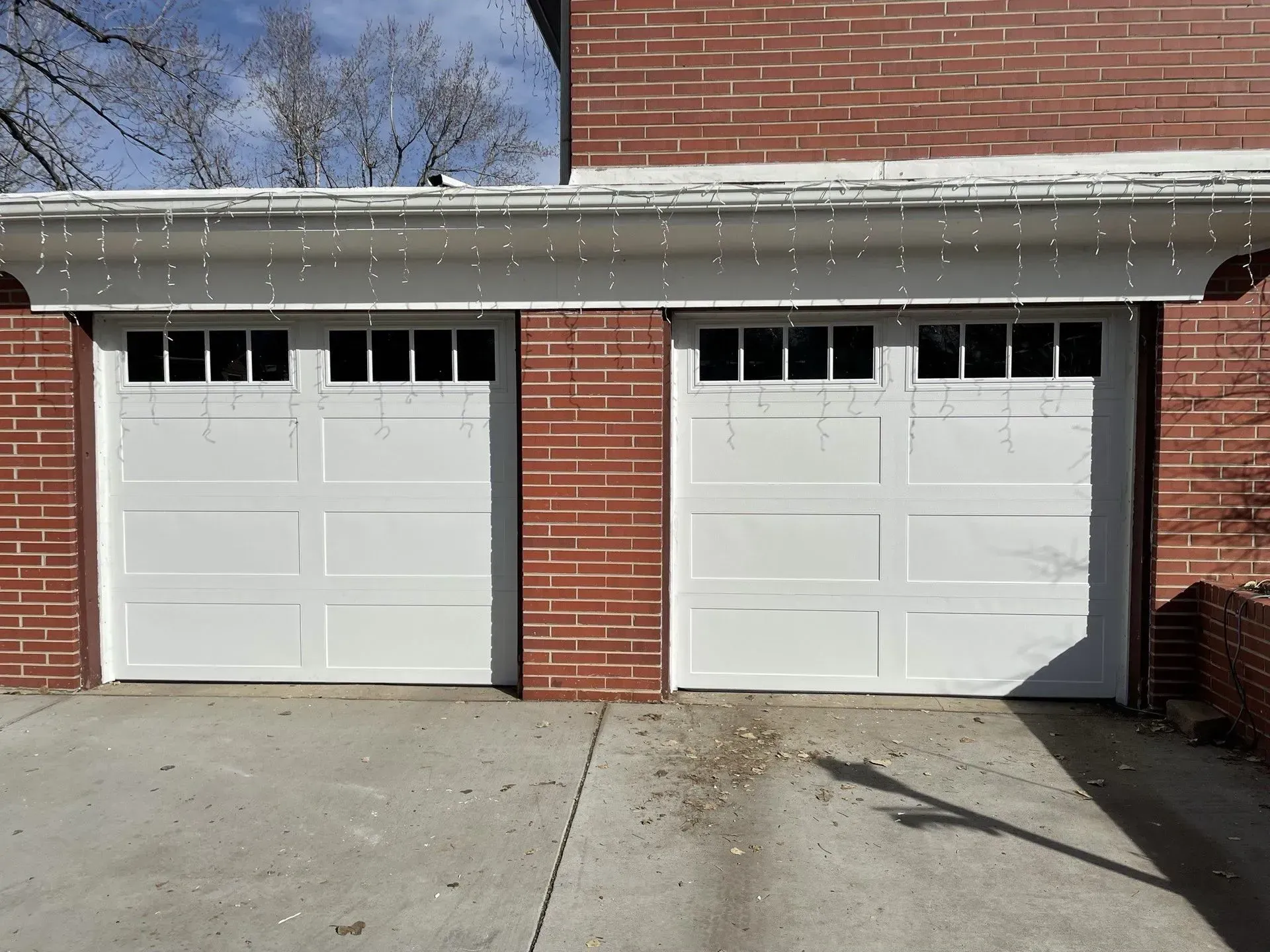 Two white garage doors on a brick house with a concrete driveway and snow on the roofline.