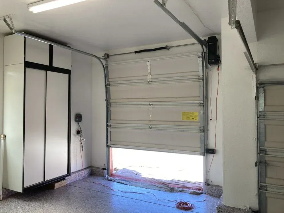 Interior of a garage with a white roll-up door, cabinets, and bright daylight near the floor opening.