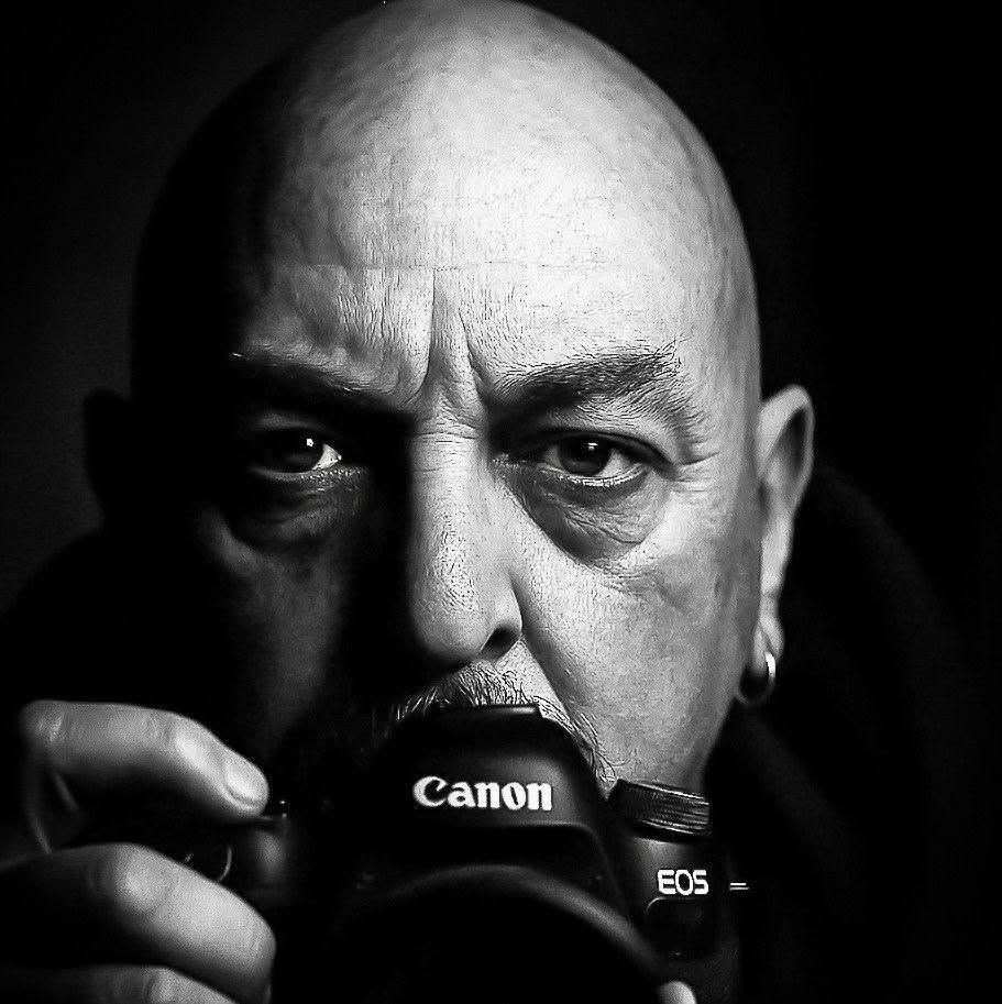 Black-and-white close-up of a bald man holding a Canon EOS camera, staring intensely at the lens