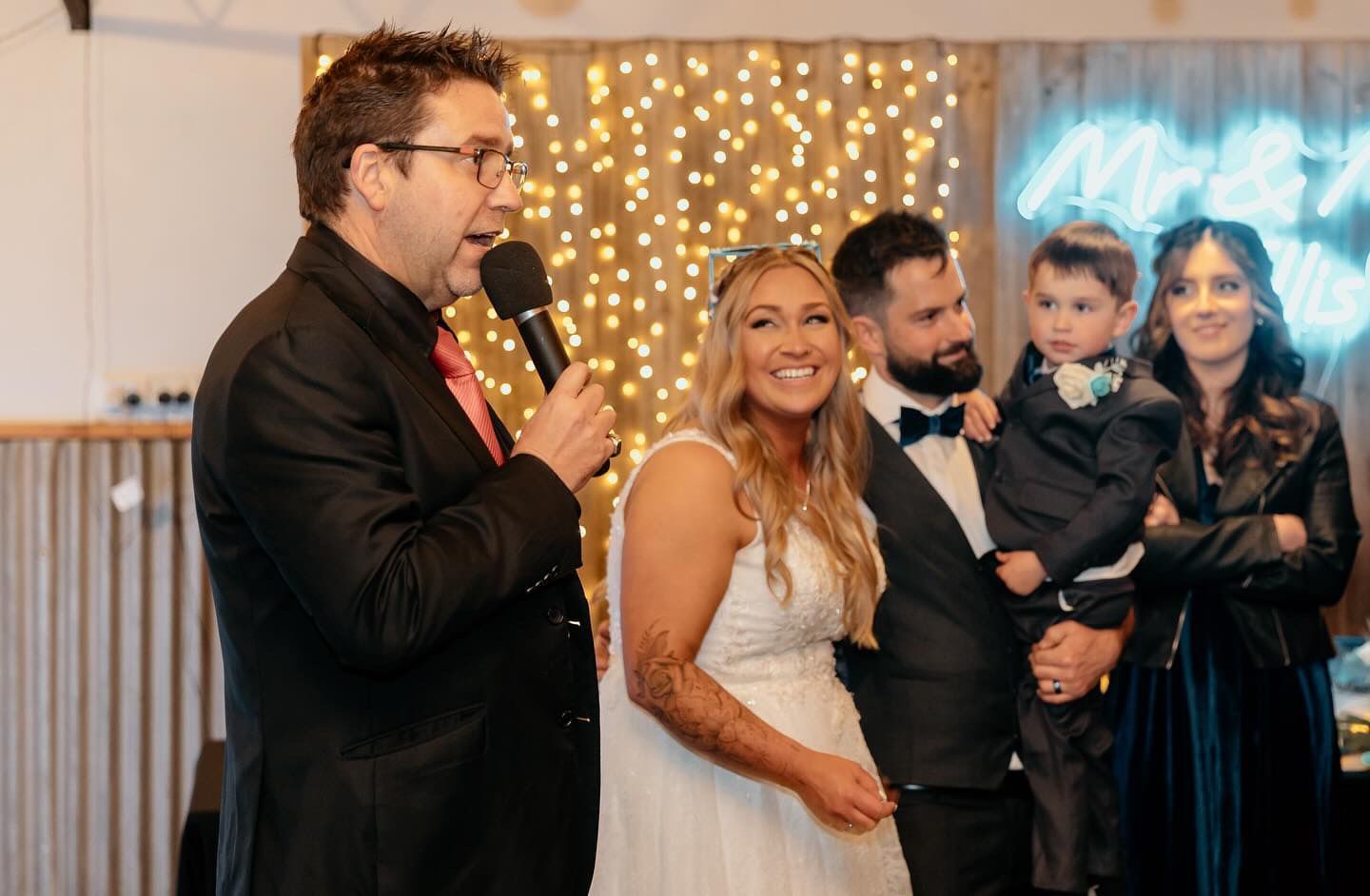 Man in a suit speaking into a microphone beside a smiling group at an indoor event with warm lights