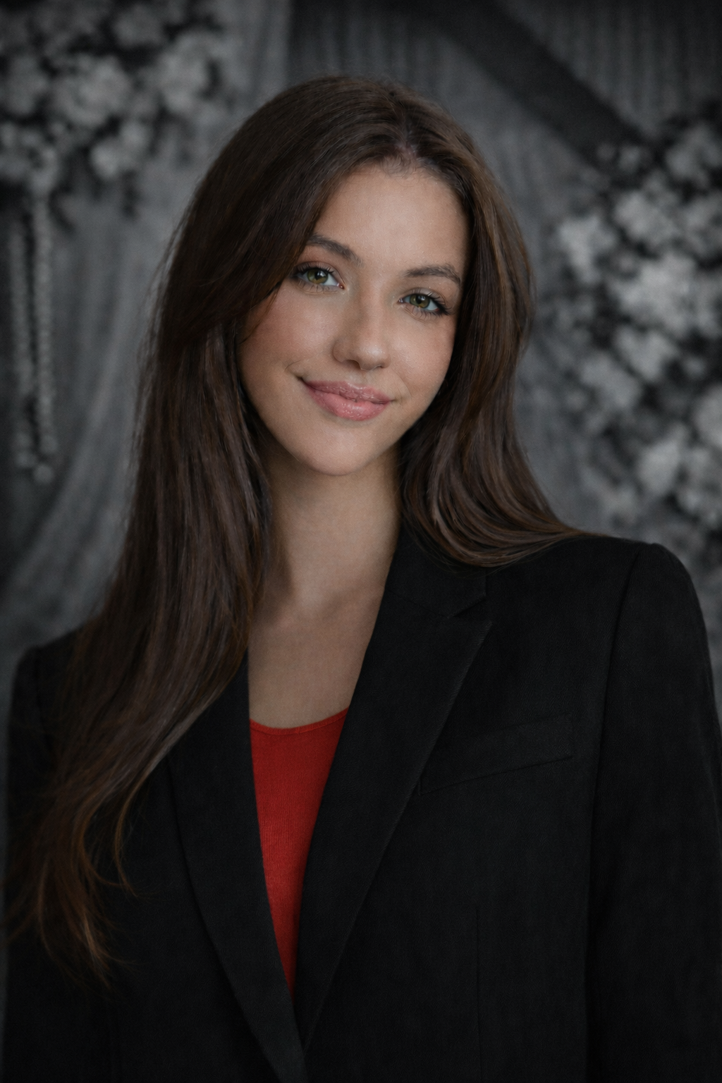 Portrait of a smiling person with long dark hair in a black blazer over a red top against a gray patterned background