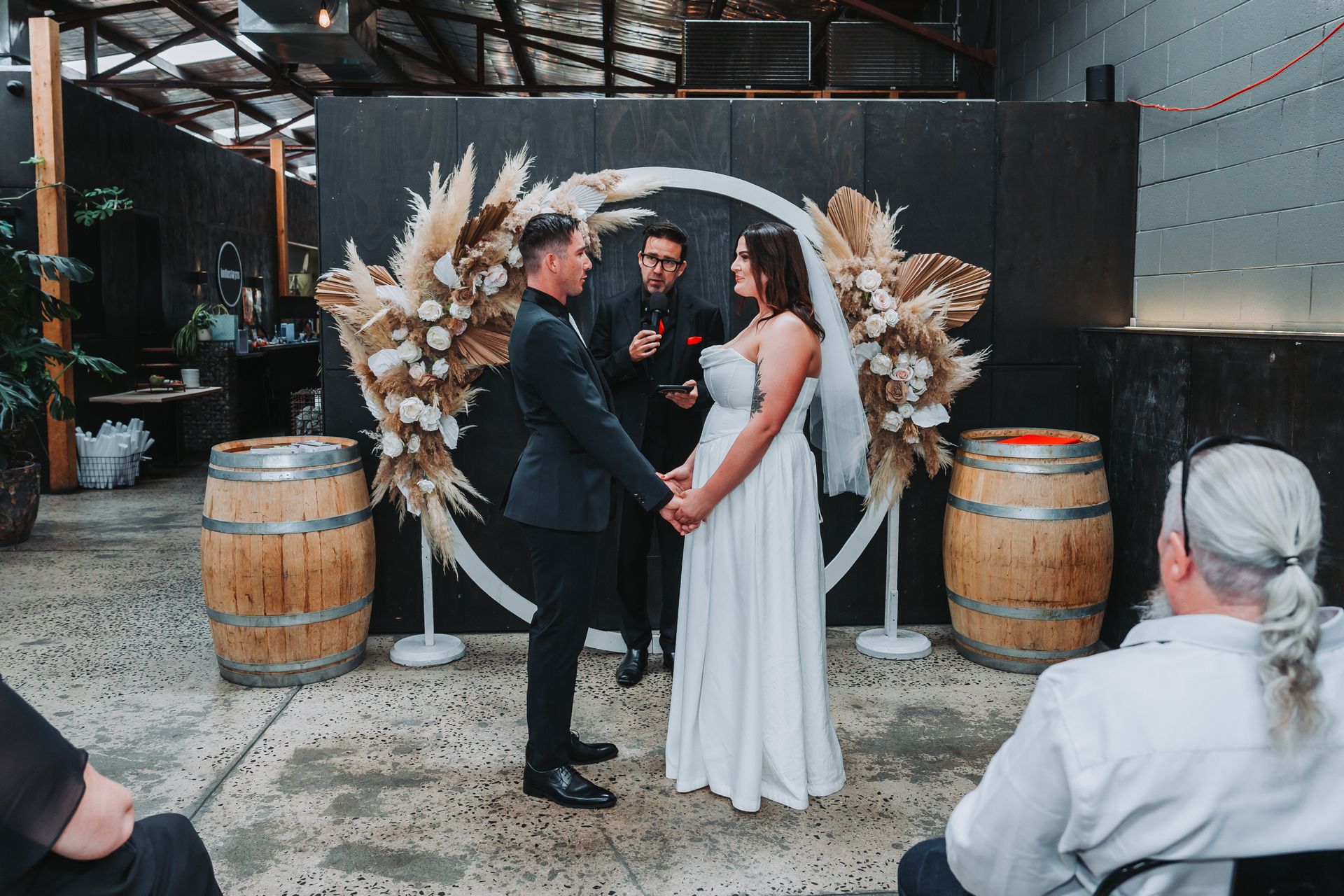 Couple exchanging vows beneath floral wings in an industrial venue, with barrels and seated guests nearby