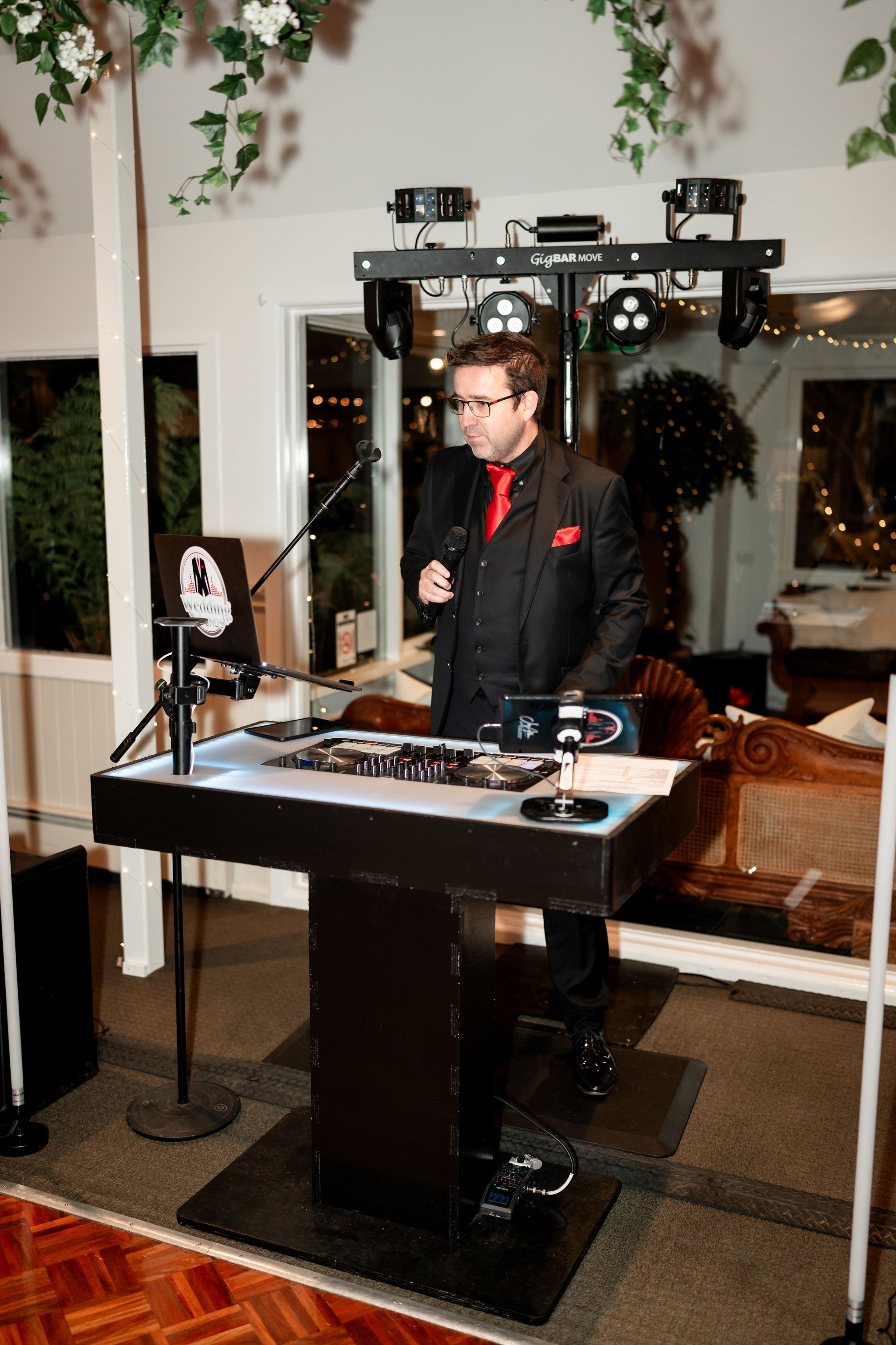 Man standing behind a DJ booth at a party, lit by stage lights with speakers and a white car in the background