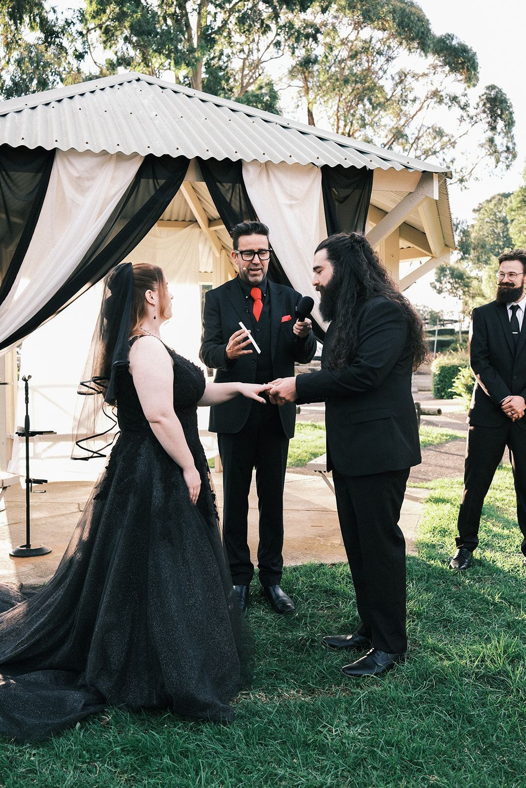 Outdoor wedding ceremony with couple exchanging rings under a gazebo, officiant in black suit nearby