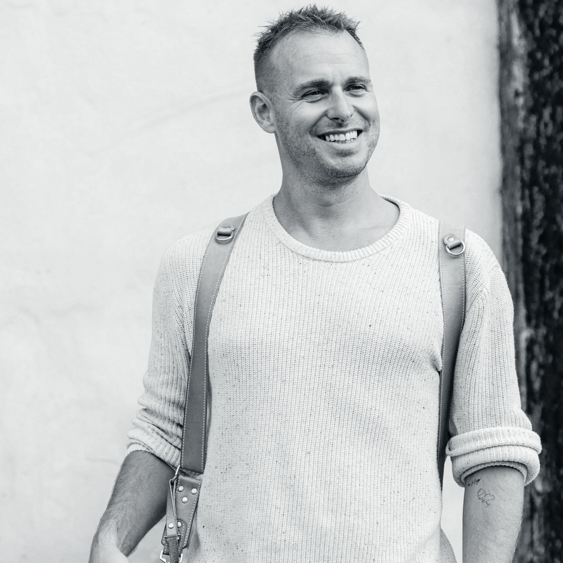 Smiling person in a light sweater with backpack straps, standing against a plain wall.