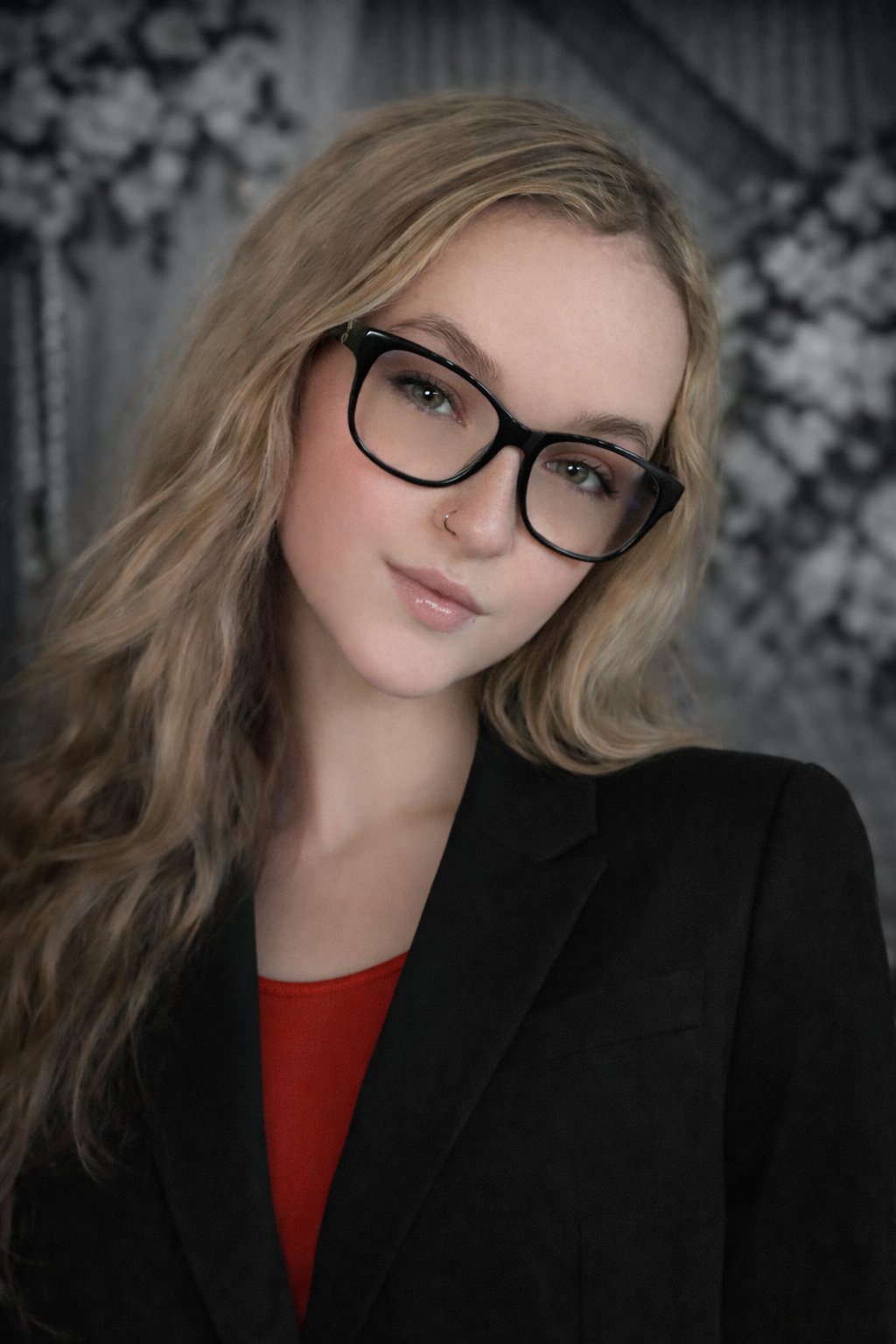 Portrait of a person with blonde hair and black glasses, wearing a black blazer over a red top.m Jackd