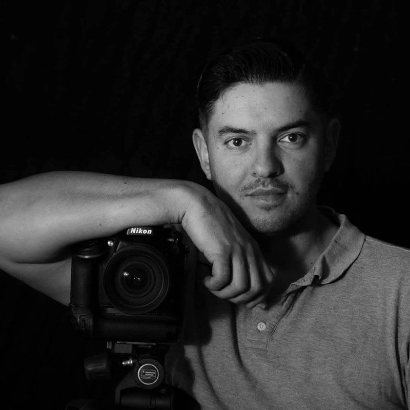 Black-and-white portrait of a man leaning on a Nikon camera against a dark background