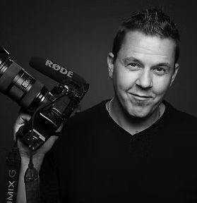 Man holding a camera rig beside a microphone, looking at the viewer in a dark studio