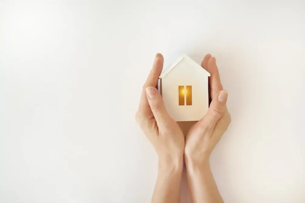 Mani che sorreggono una piccola casa bianca con una finestra luminosa su uno sfondo chiaro.