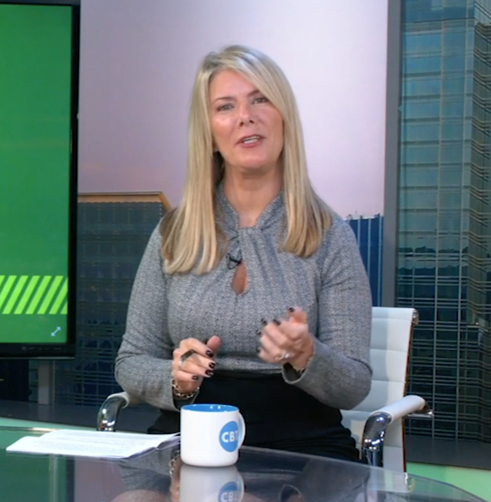 Woman in gray top and black skirt, seated at a desk, speaking in a studio setting with cityscape backdrop.