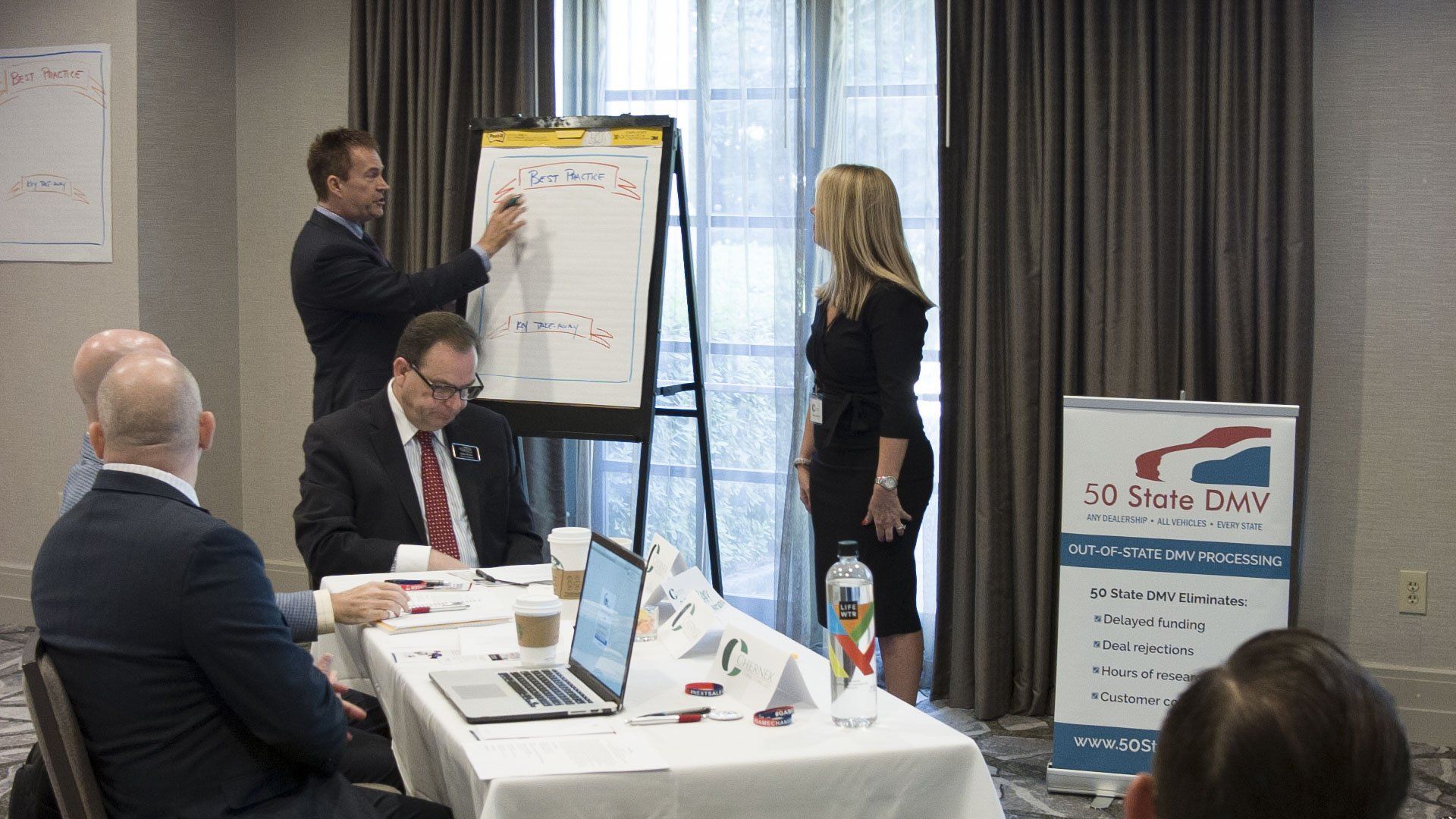 A man presents at a whiteboard in a meeting; others watch. A banner and laptop are also present.