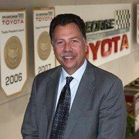 Man in suit smiling, posing in front of a Toyota dealership sign.