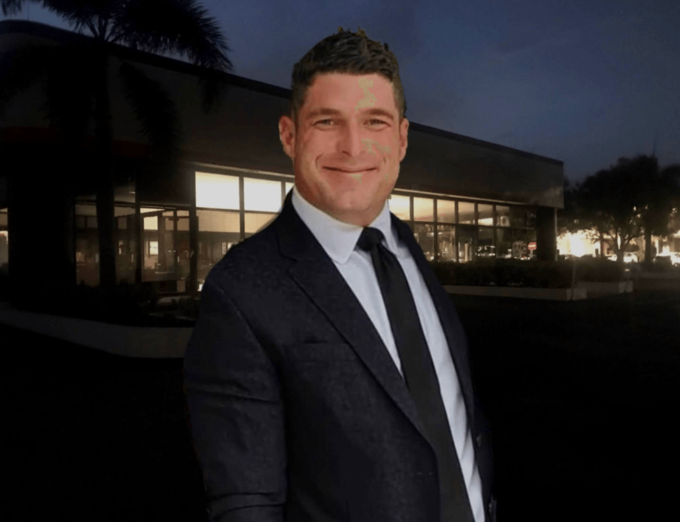 Man in suit smiles in front of a building at dusk.