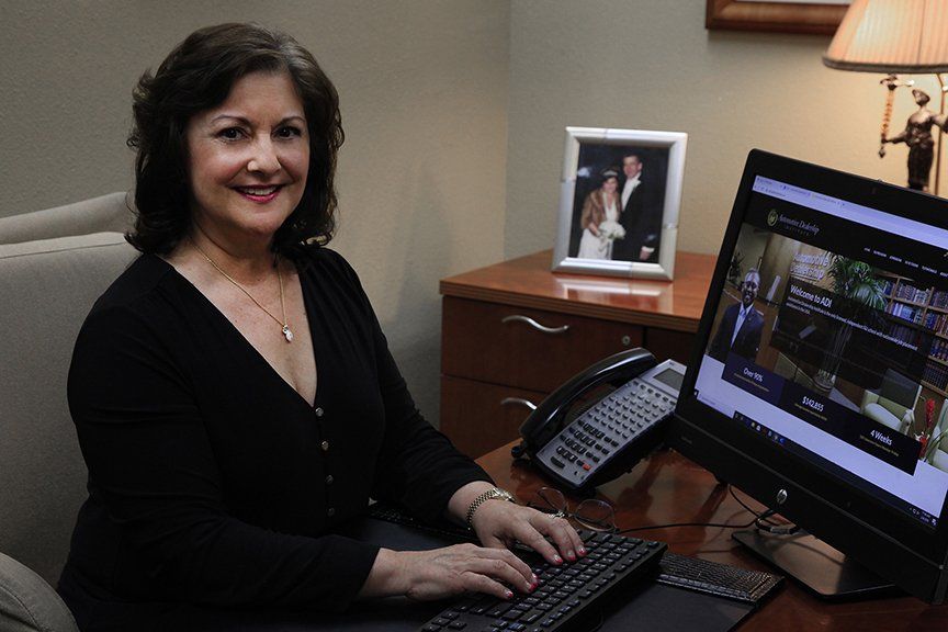 Woman sitting at a desk, typing on a keyboard, smiling. There is a computer and photo on desk. Office setting.