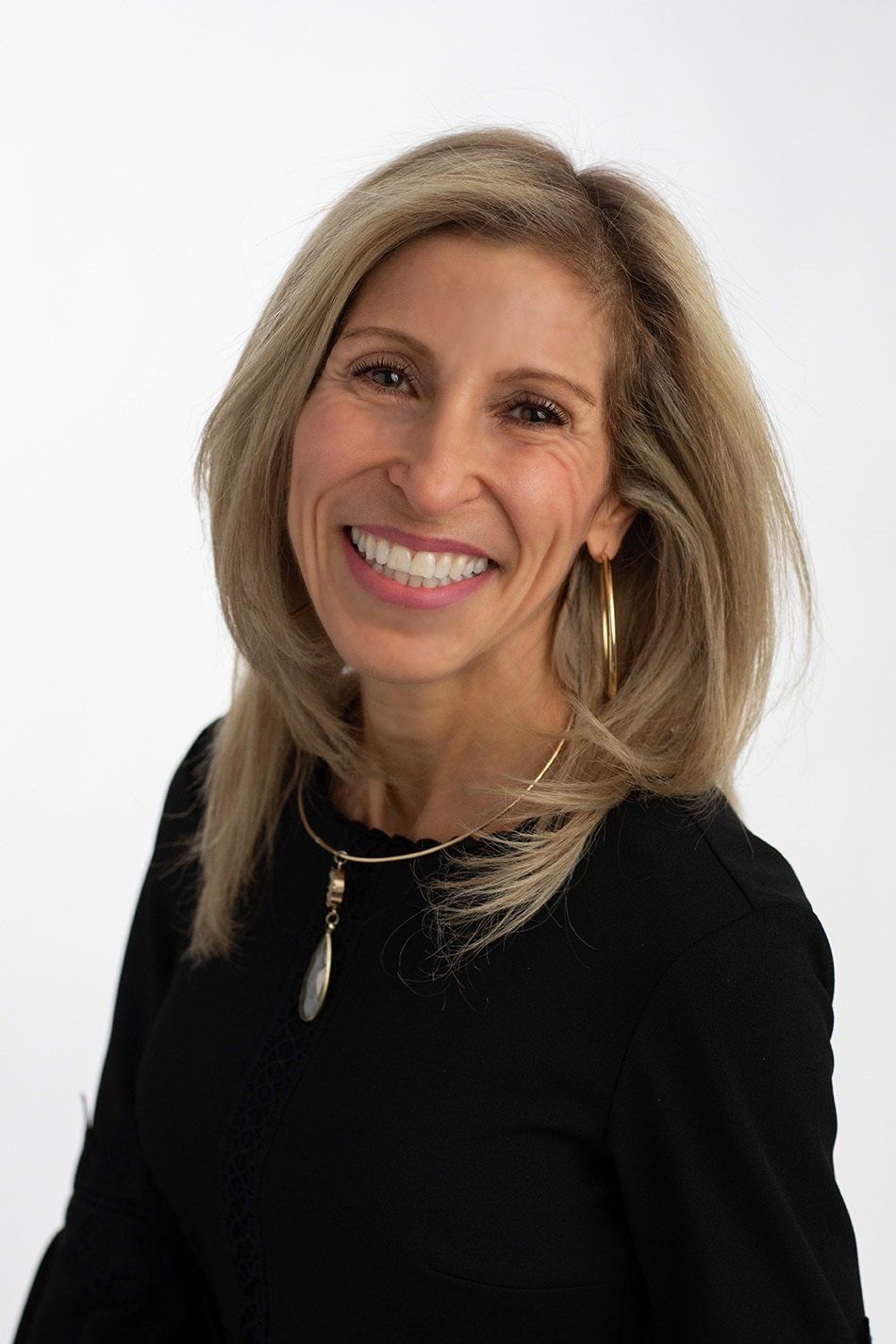 Woman with blond hair, smiling, wearing a black top and necklace, posing against a white background.