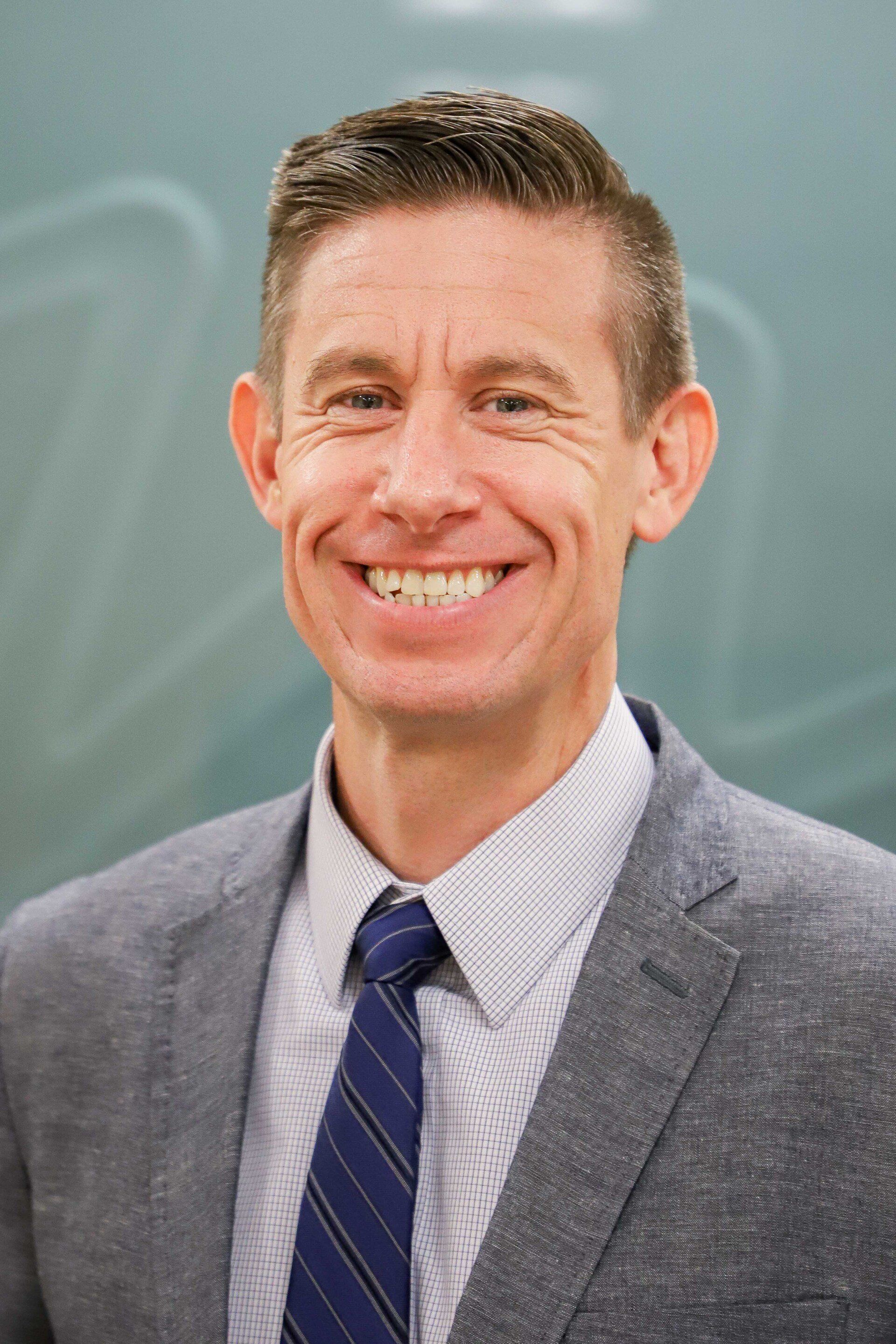 Man in gray blazer and blue tie smiles in front of a chalkboard.