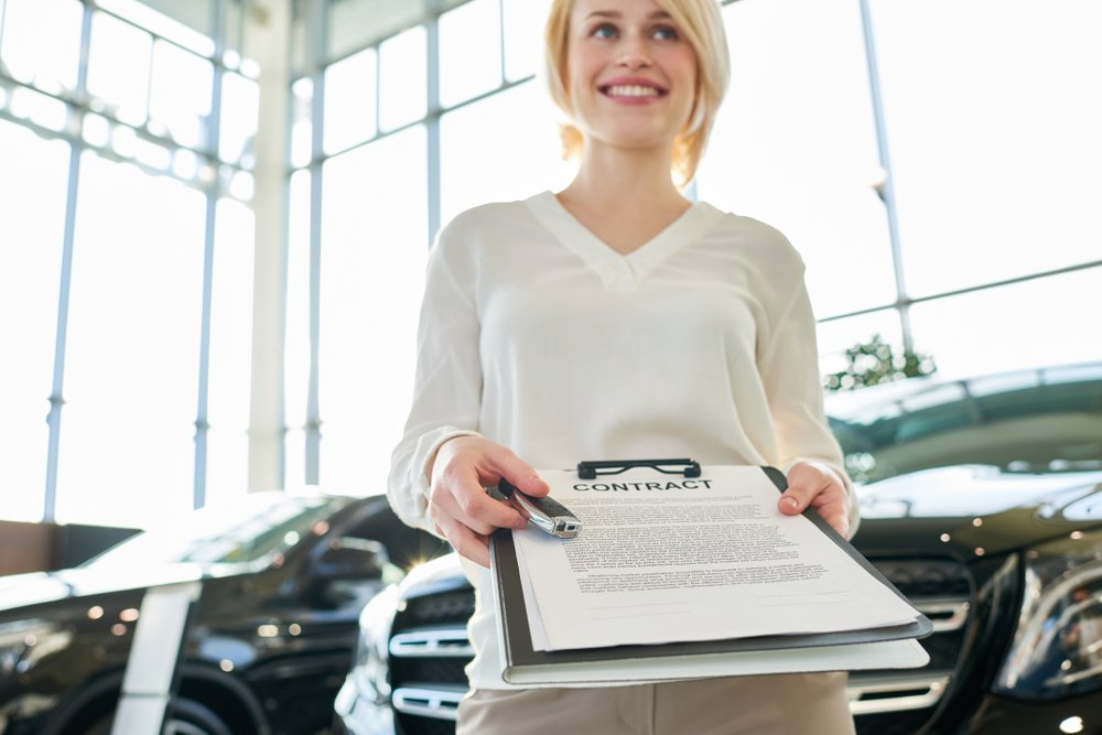 Woman in car dealership holds car keys and contract, smiling.