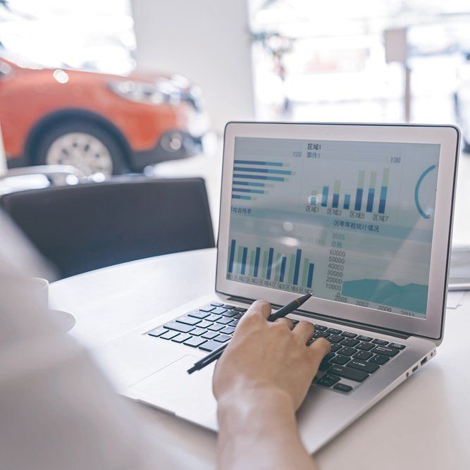 Person working on a laptop with charts in a car dealership; a car is in the background.