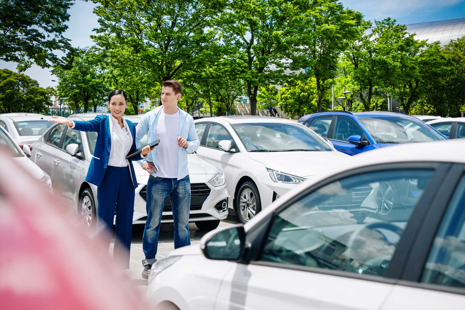 Woman in suit gestures, showing cars to a man in a car lot with other cars and trees.