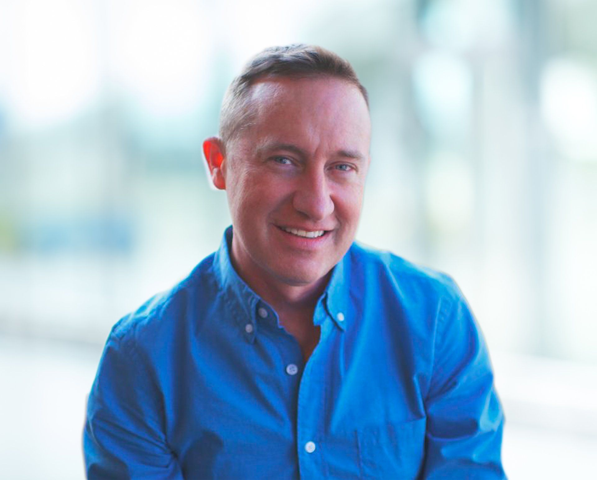 Man with short hair smiling, wearing a blue button-down shirt; soft, blurred background.