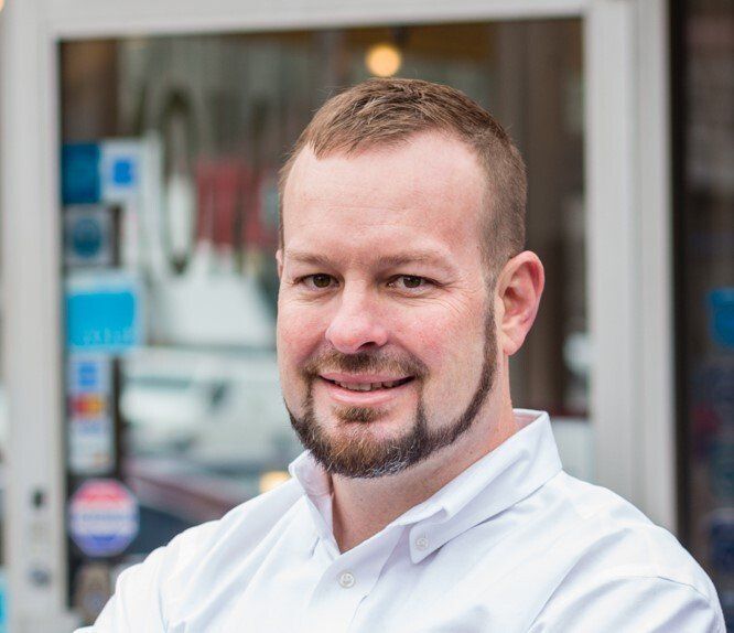 Man with short brown hair and beard smiles in front of a storefront.
