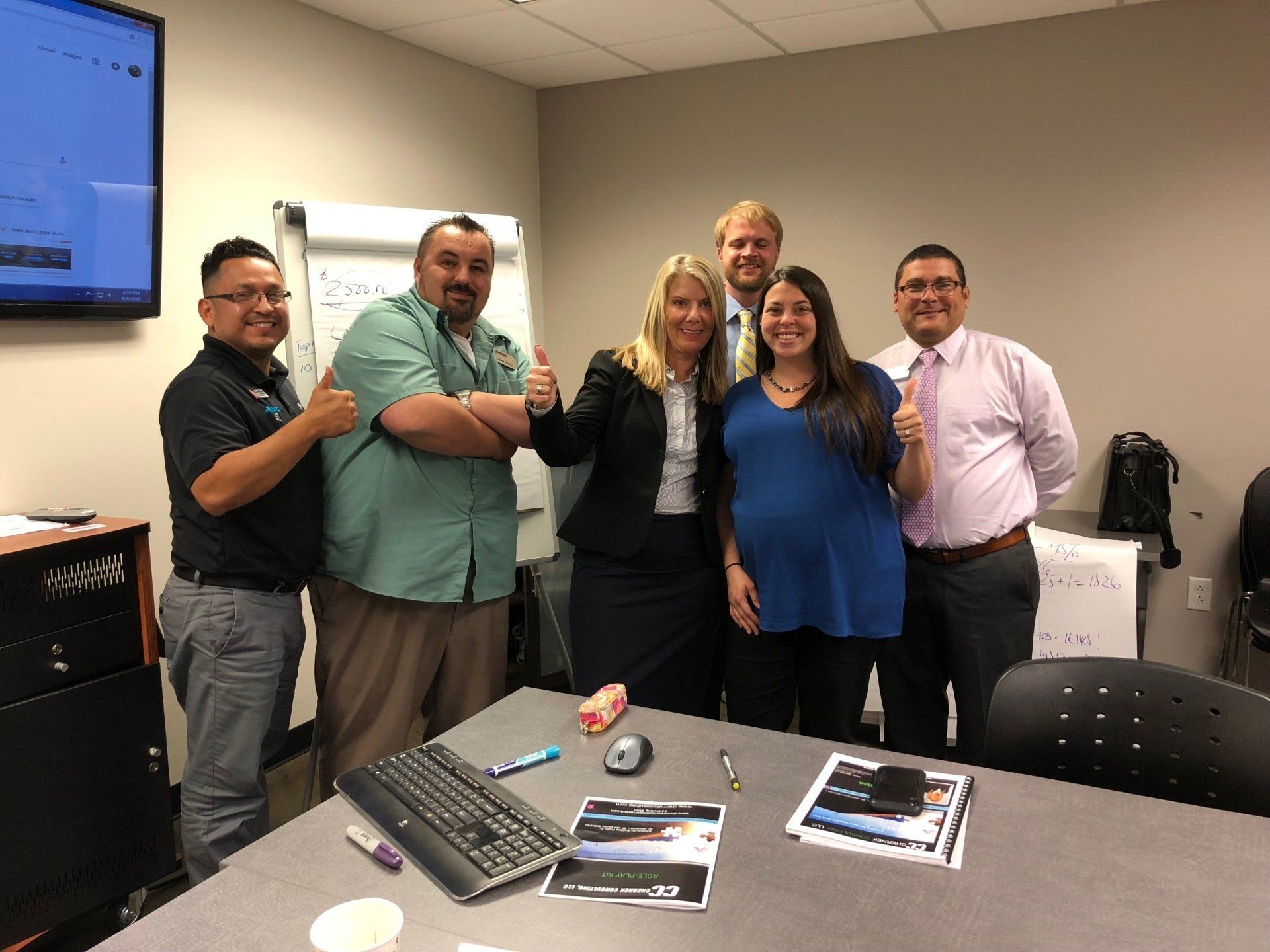 Group of six people giving thumbs up in an office setting, smiling and happy.