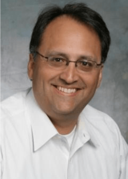 Man with glasses smiles, wearing a white button-down shirt. Blurred, neutral background.