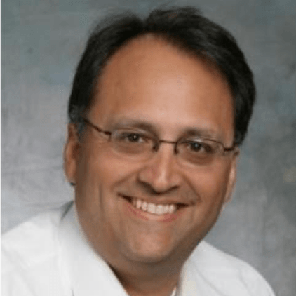 Man wearing glasses, smiling, in a white shirt, against a soft gray background.