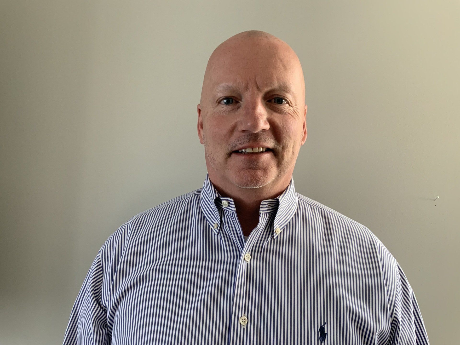 Man with bald head smiles, wearing a striped blue and white shirt. Plain wall background.