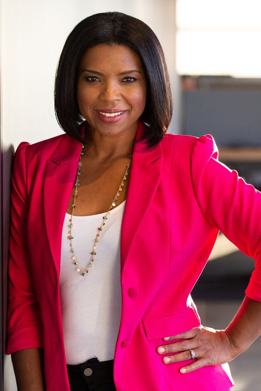 Woman in pink blazer smiles, leans against wall, wearing necklace, arm on hip.