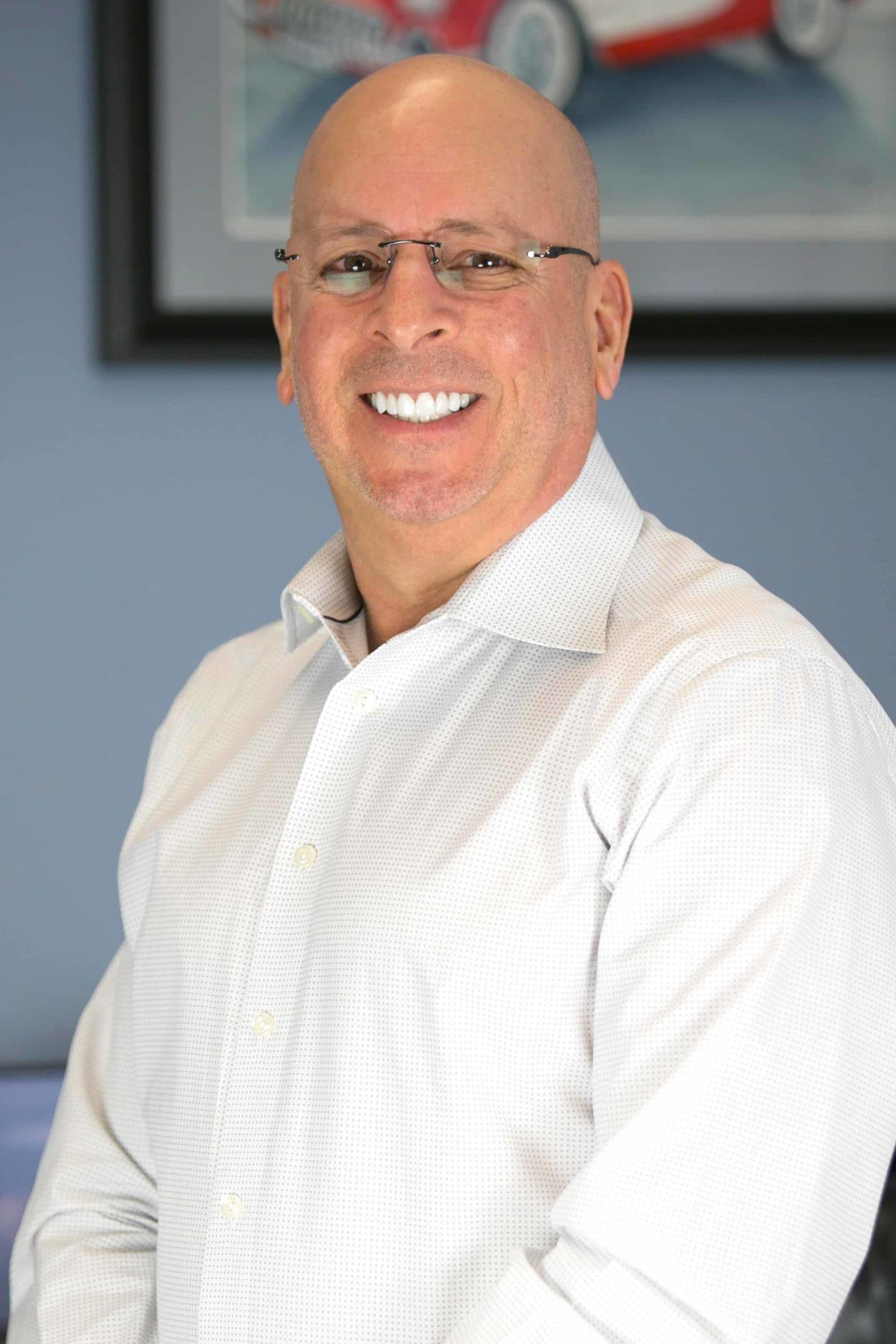 Bald man wearing glasses smiling, in white button-up shirt; blue wall with framed image in background.