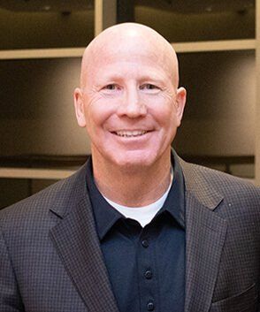 Bald man smiles, wearing a dark suit jacket over a black shirt and white undershirt. Neutral background.