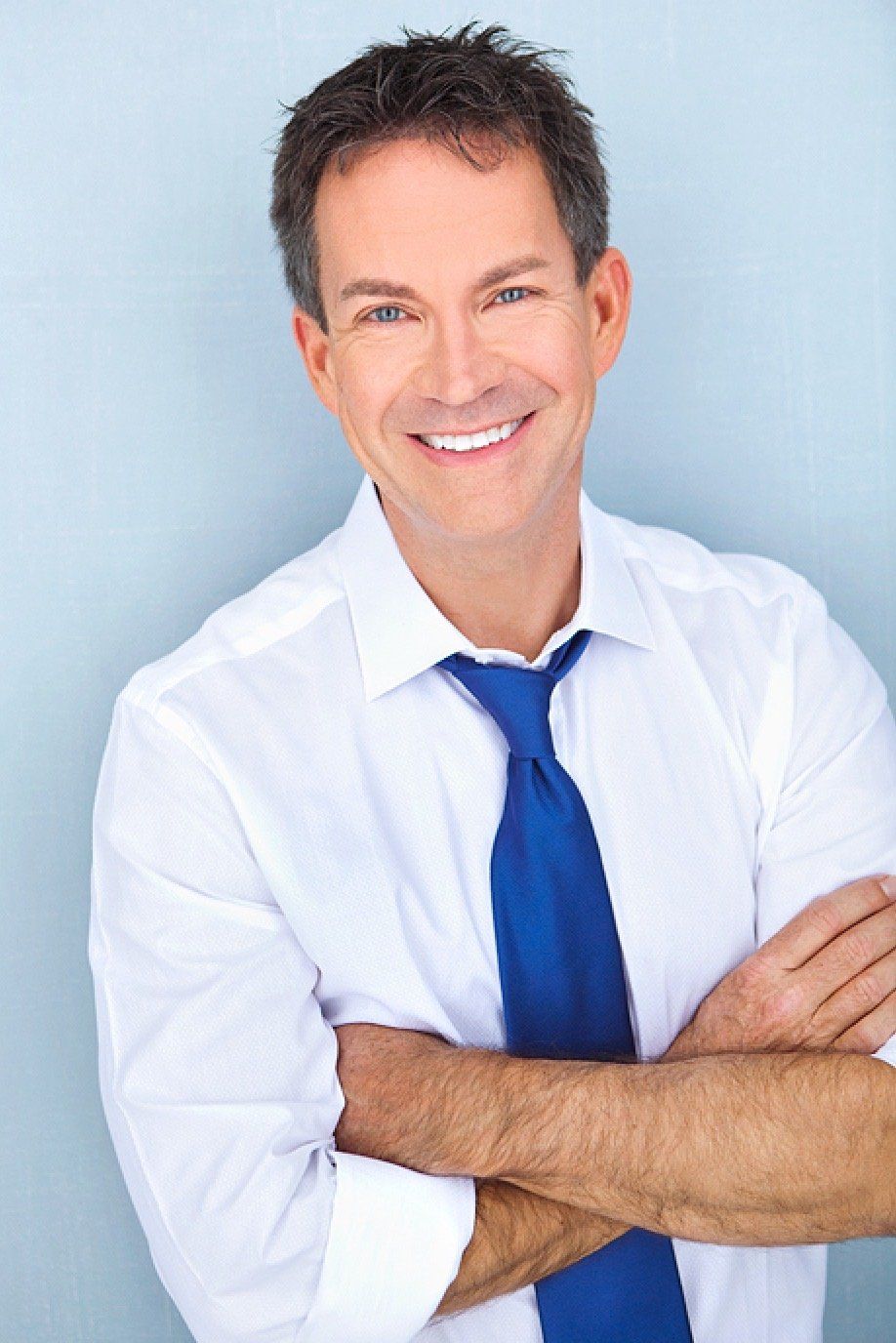 Man in white shirt and blue tie smiles, arms crossed against a light blue background.